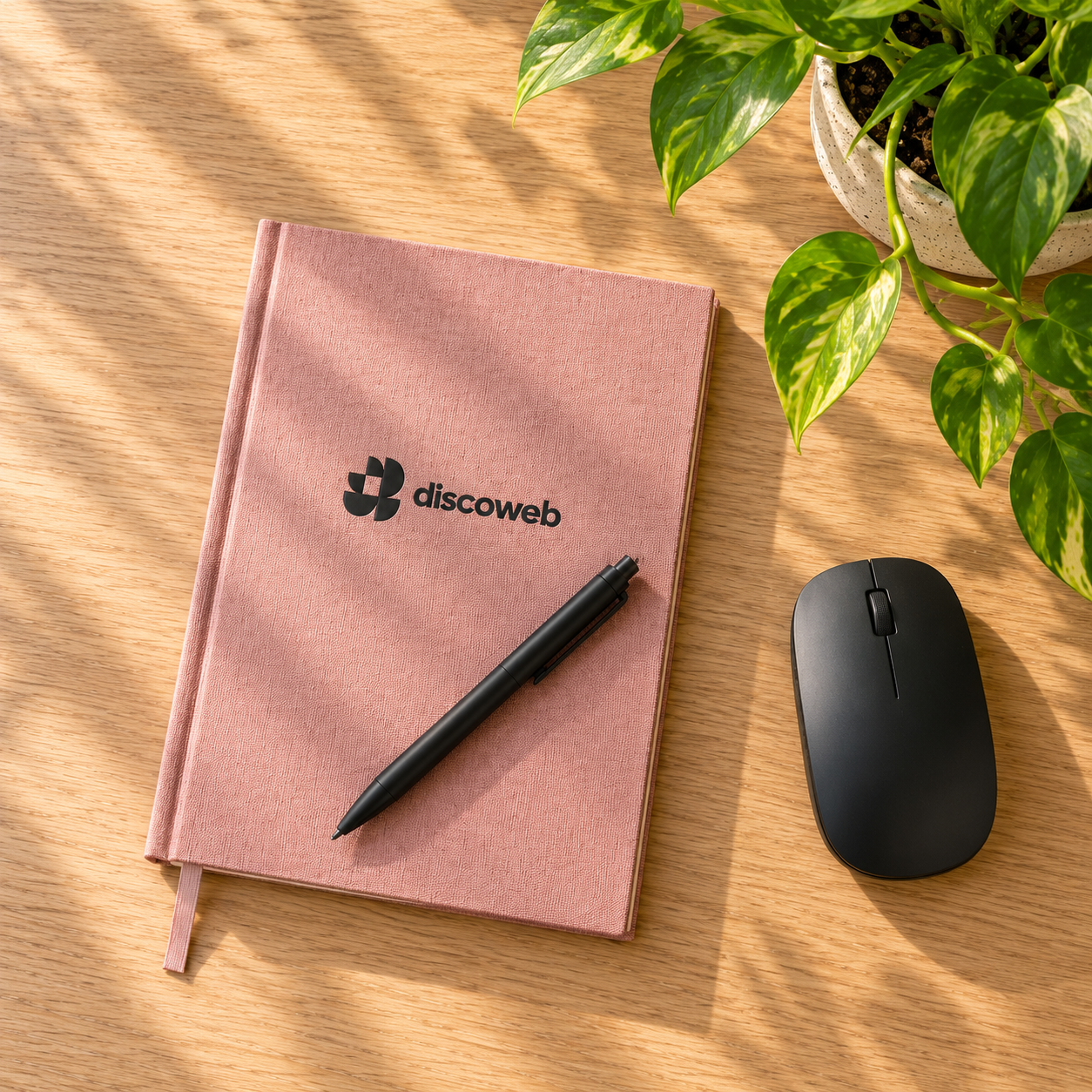 A dusty pink discoweb notebook and pen on a sunlit wooden desk beside a mouse and a trailing pothos plant
